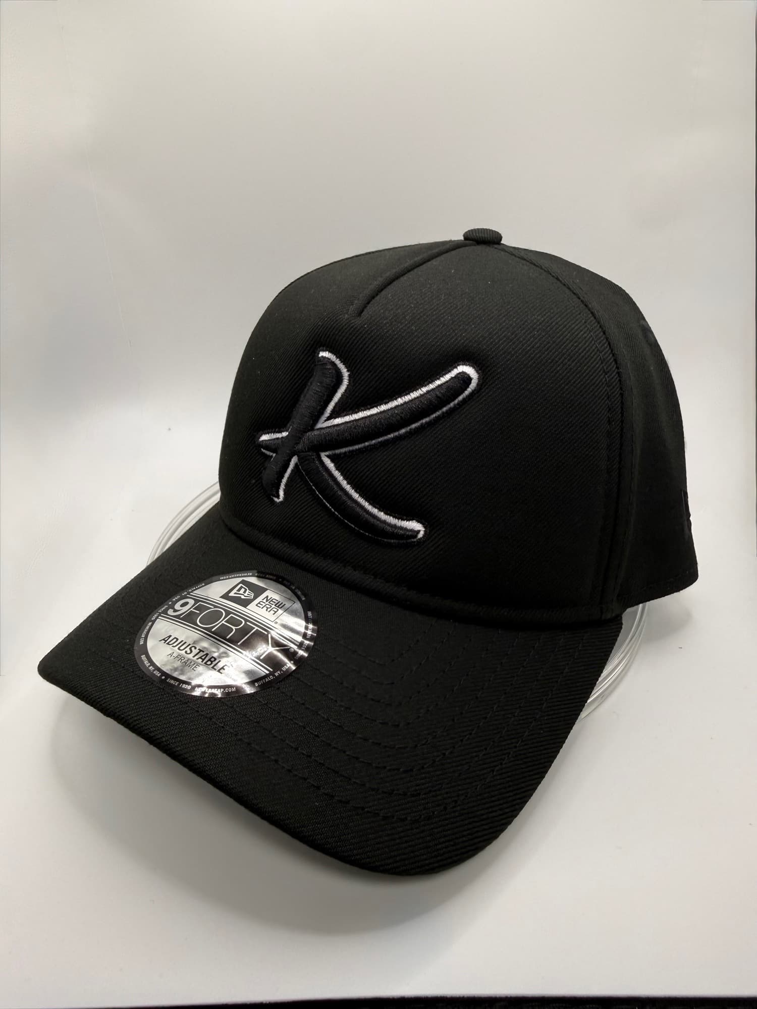 Black New Era 9Forty cap with a script letter K embroidered in black with a white outline